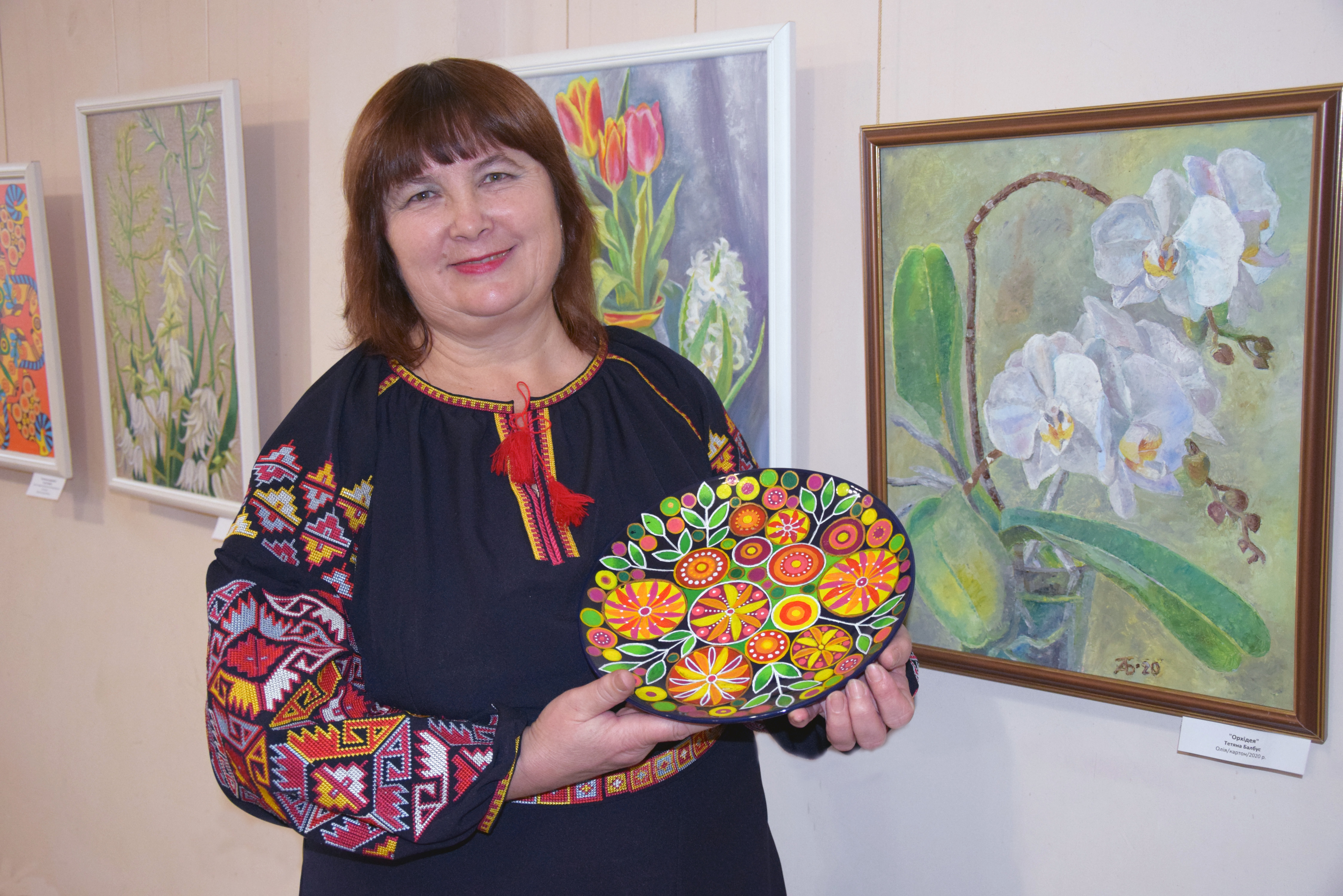 Керівниця гурту «Палітра» Тетяна Балбус на тлі своїх картин демонструє ще одну авторську роботу — декоративно розписану керамічну тарілку. Фото автора